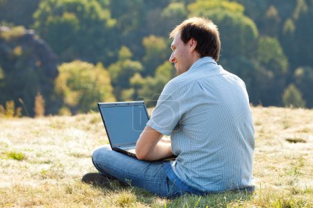 jeune homme utilisant un ordinateur portable assis sur l'herbe sur la colline 